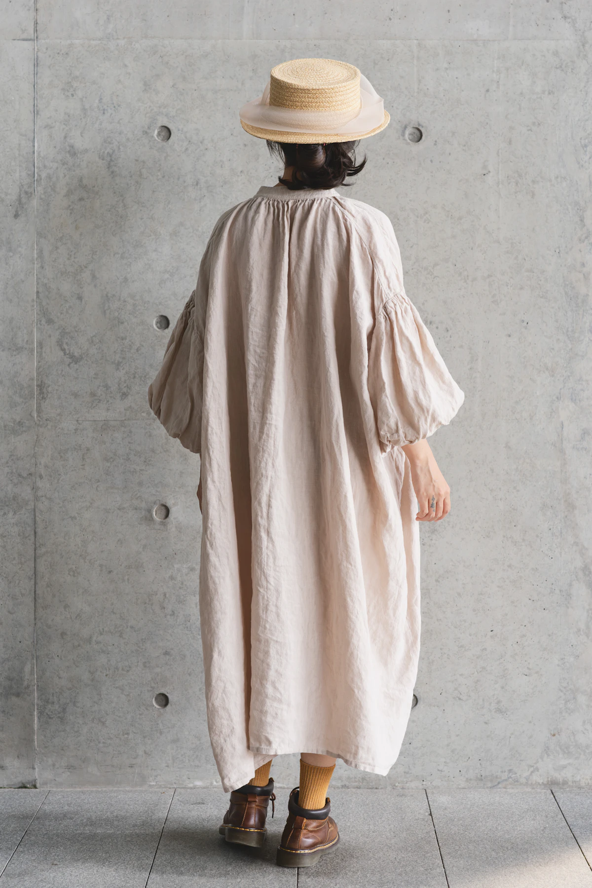 A woman in a straw hat wearing the Haveli maxi dress in oat linen, seen from behind against a concrete wall
